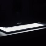 A dramatic, low-light image of a slightly open laptop with glowing keys on a dark desk.